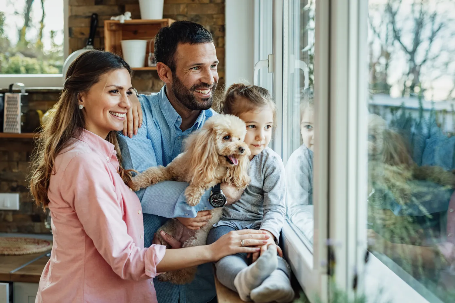 HanseMerkur Versicherung - Tierversicherung - eine lächelnde Familie mit ihrem Pudel am Fenster.