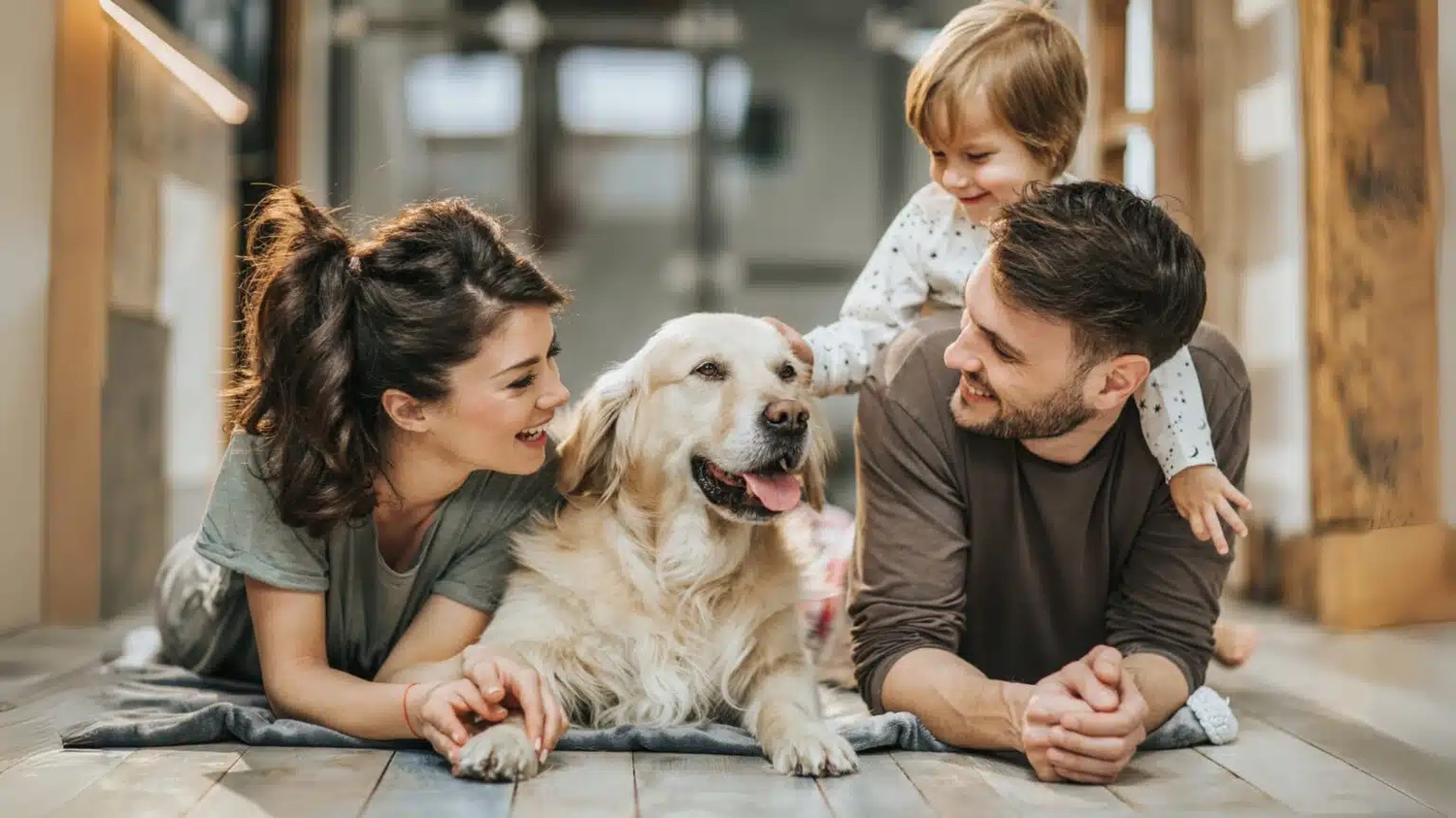 HanseMerkur Versicherung - Tierversicherung - Eine Familie mit einem kleinen Kind lächelt, während sie auf dem Boden liegt und ihren Golden Retriever streichelt.